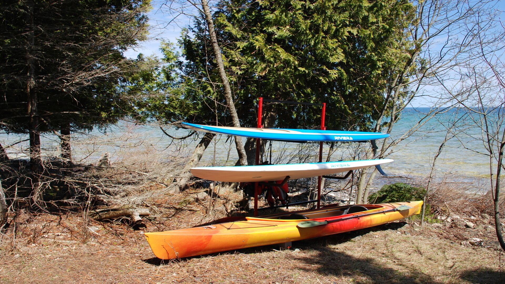 A row of kayaks and a canoe on a rocky shore with trees and a body of water in the background.