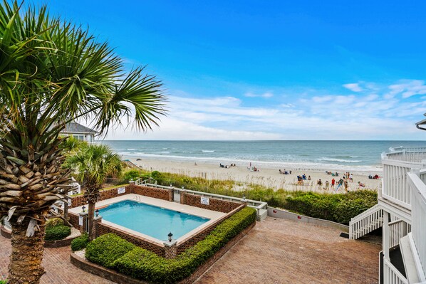 Relax by the pool surrounded by lush greenery and a brick patio overlooking the beach.