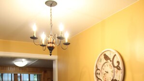 A dining room with a wooden table set for six, surrounded by chairs. A chandelier hangs from the ceiling, and a decorative plate is mounted on the wall. - Beautiful Views, Sunrises, Dog Friendly, Comfortable! (Penn Yan)