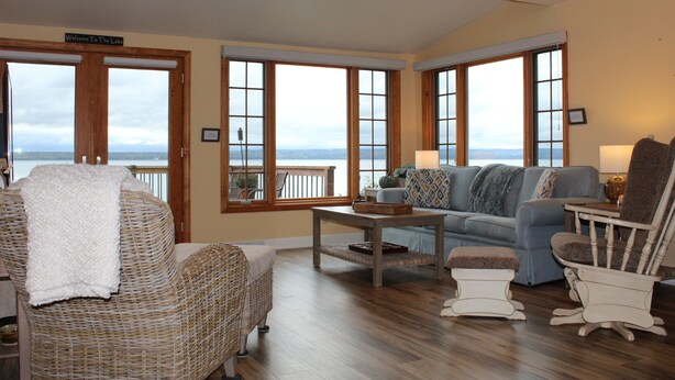 A spacious living room with a view of the lake, featuring a wicker chair, a blue sofa, and a wooden coffee table. - Beautiful Views, Sunrises, Dog Friendly, Comfortable! (Penn Yan)