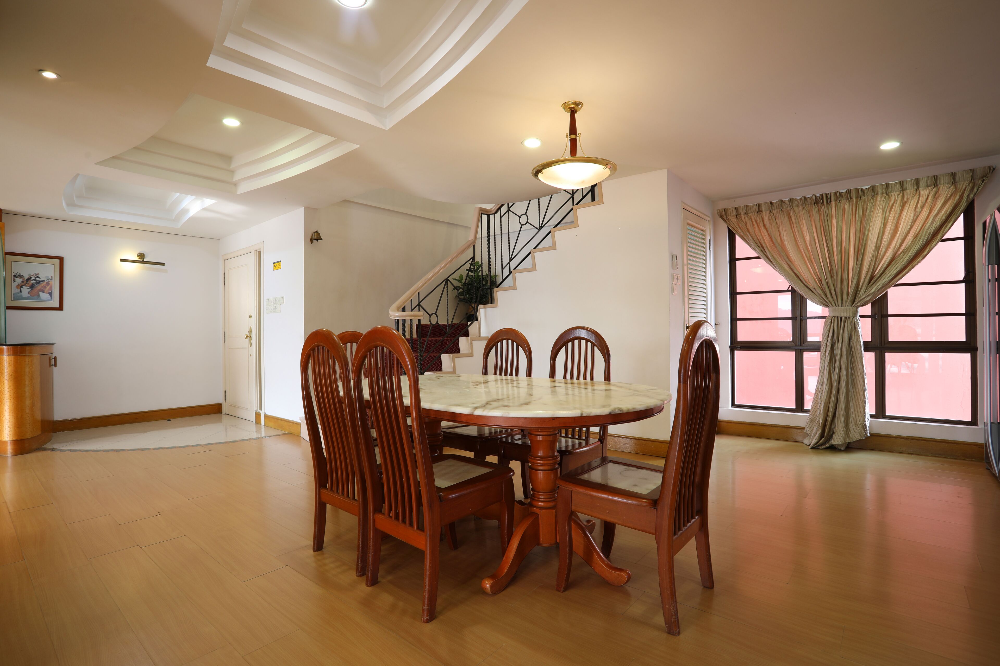 A dining area with a round table and wooden chairs, a staircase with a railing, and a window with curtains.