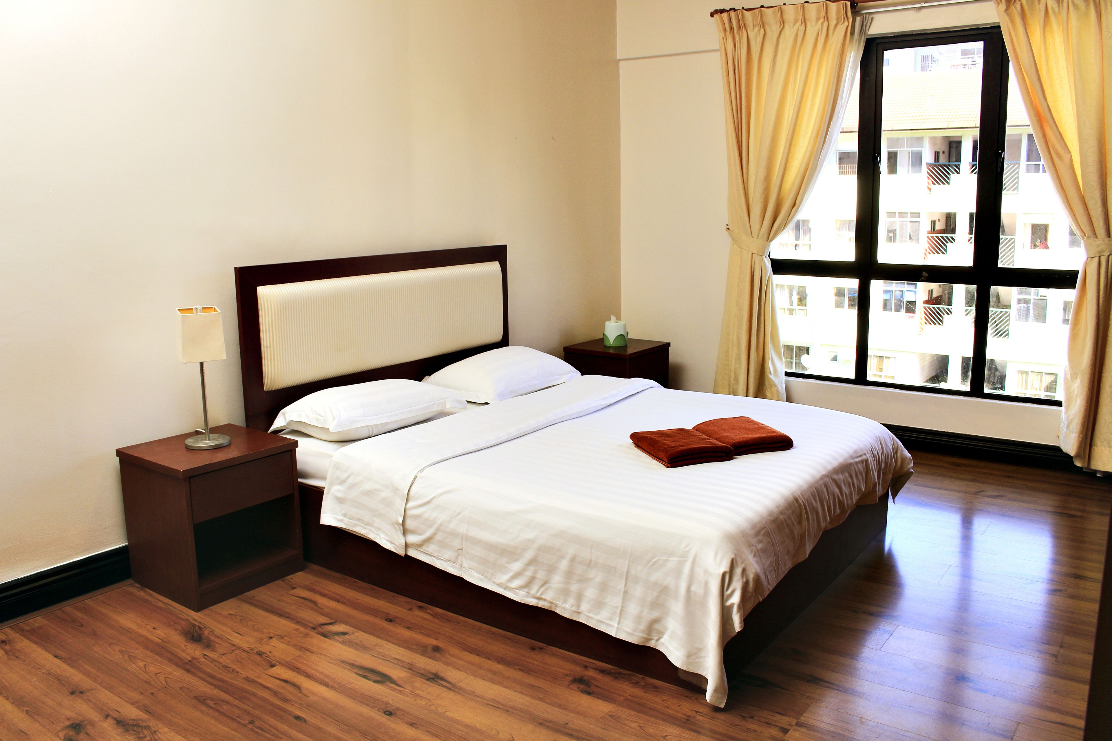 A neatly made bed with white linens, a wooden nightstand with a lamp, and a window with curtains.