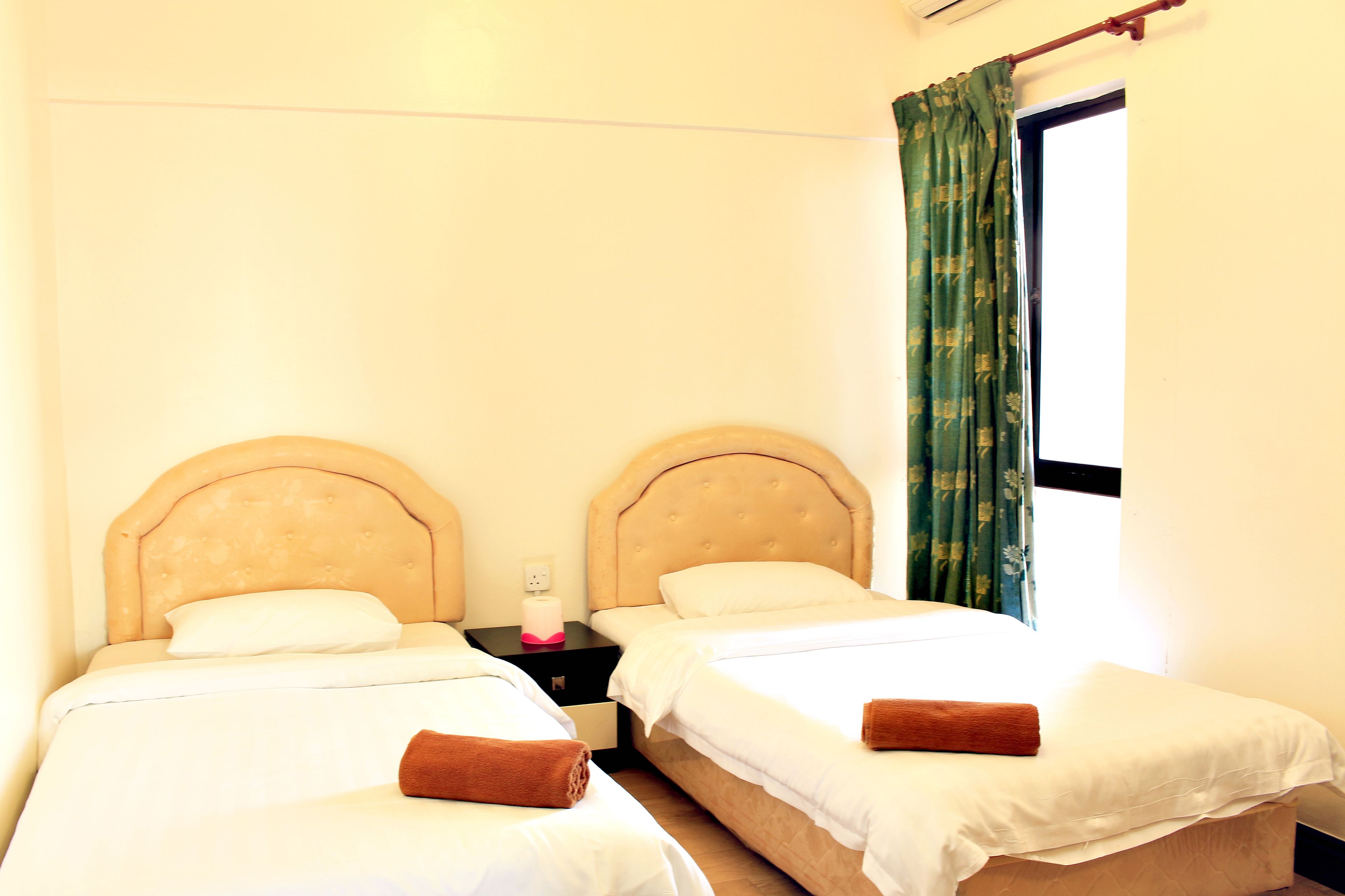 A room with two beds, each with a headboard, a nightstand with a lamp, and a window with curtains.