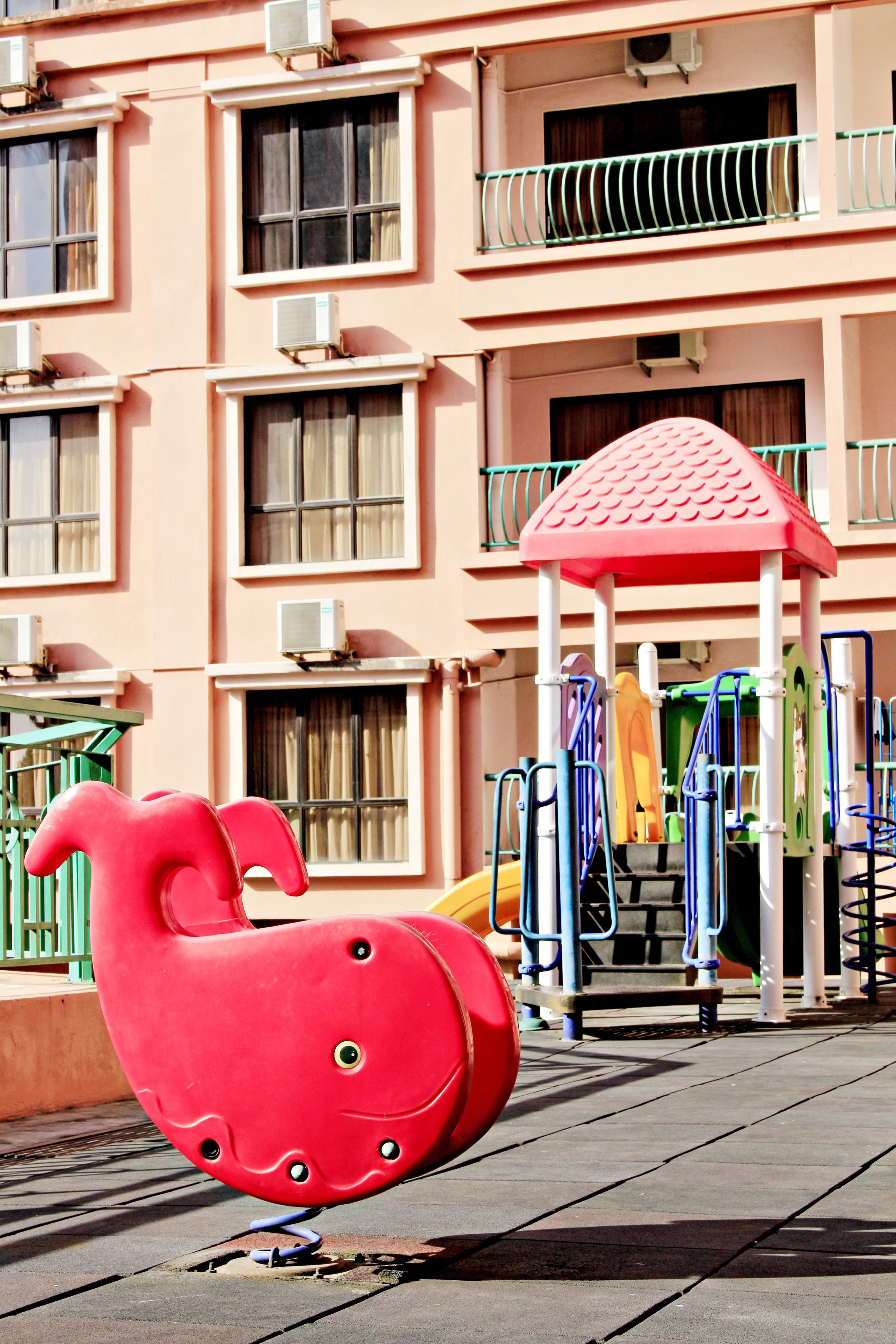 A playground with a red spiral slide and a pink building with multiple windows and balconies in the background.