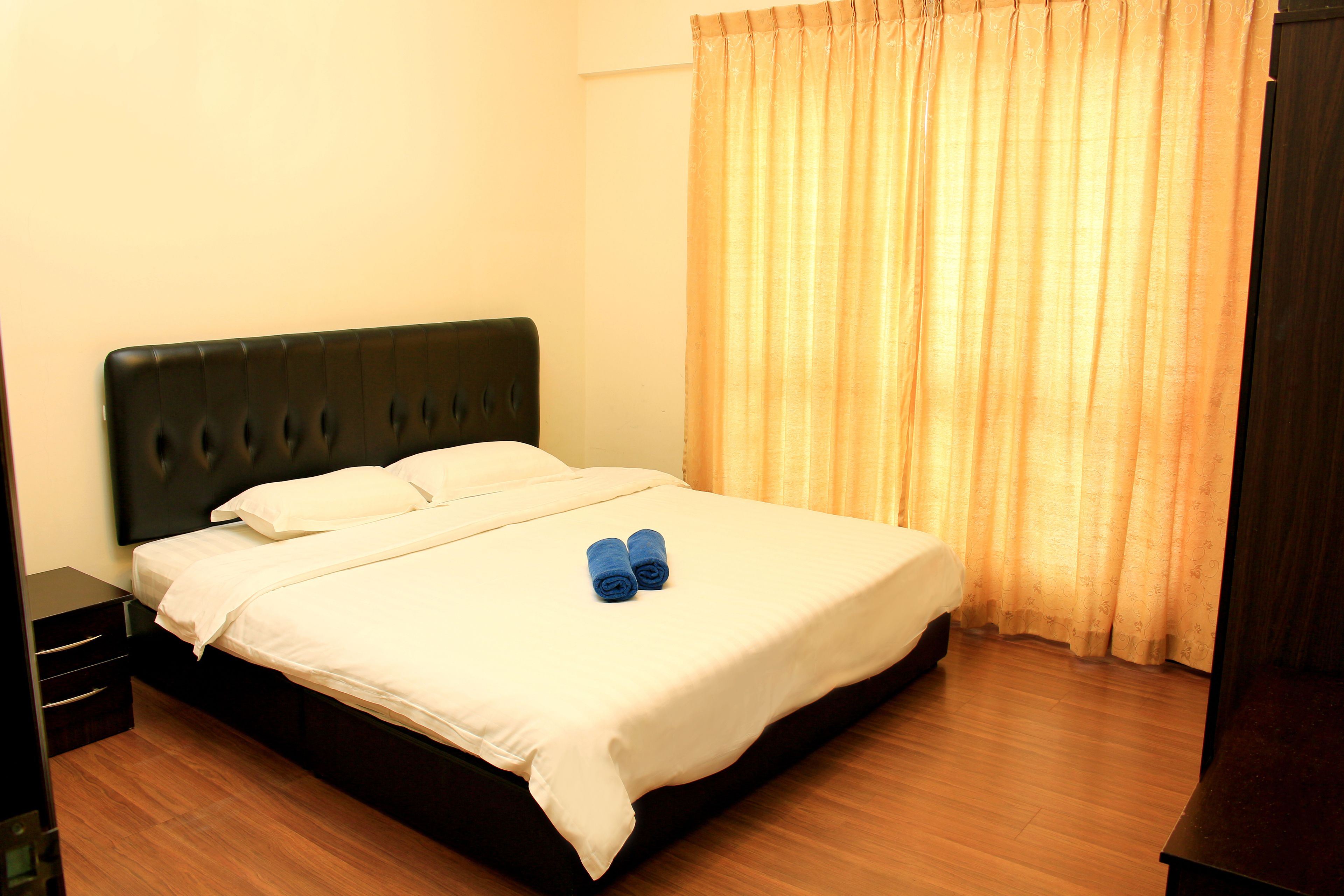 A neatly made bed with white linens and two rolled towels, a dark headboard, and a wooden floor.