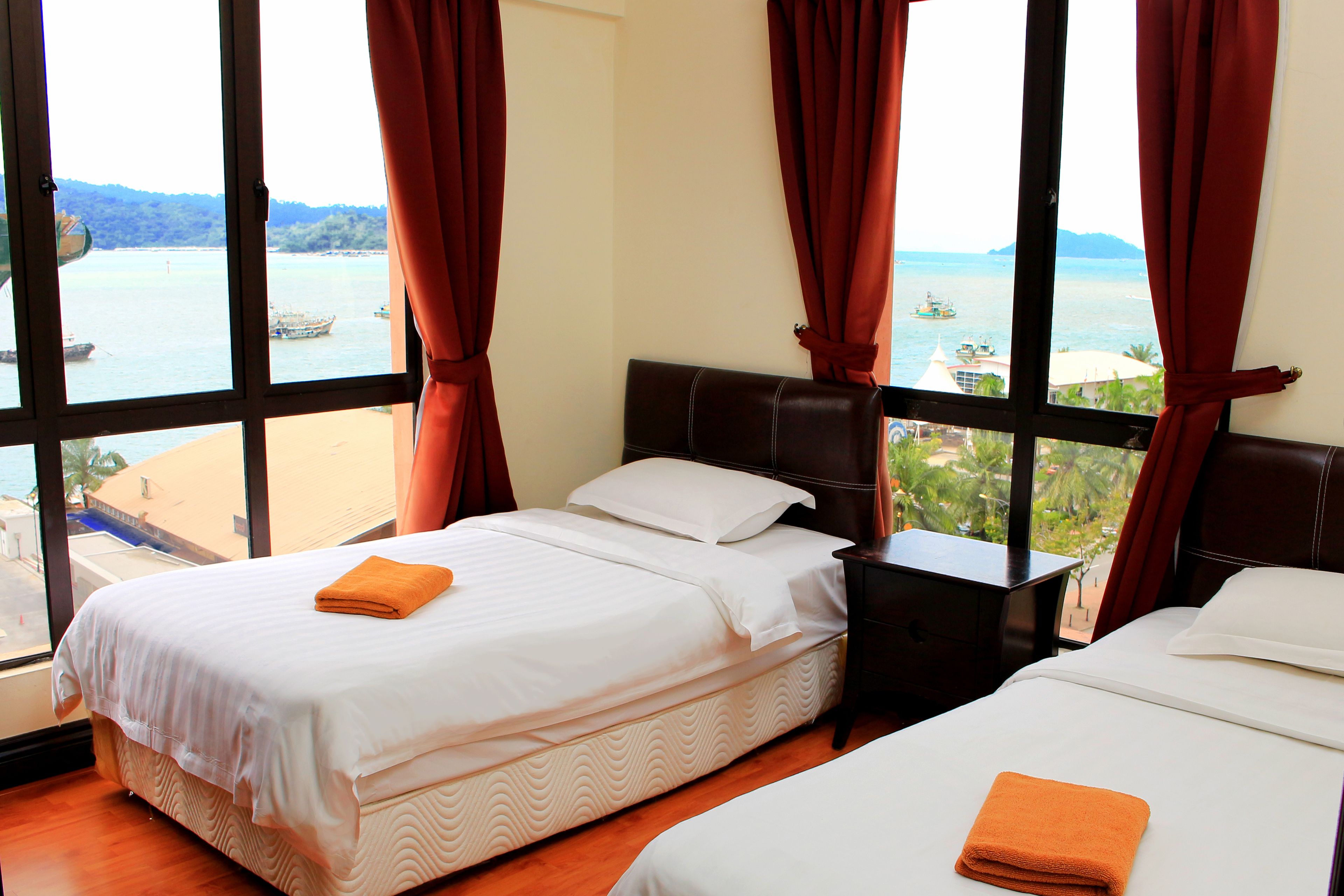 Habitación de hotel con dos camas, ventanas amplias con cortinas rojas y vista al mar y a los barcos.