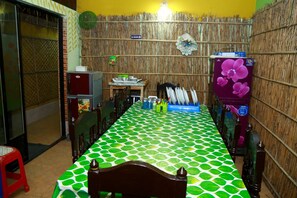 Uma área de refeições com toalha de mesa estampada em verde e branco, uma geladeira com desenho de flor roxa e uma parede de bambu.