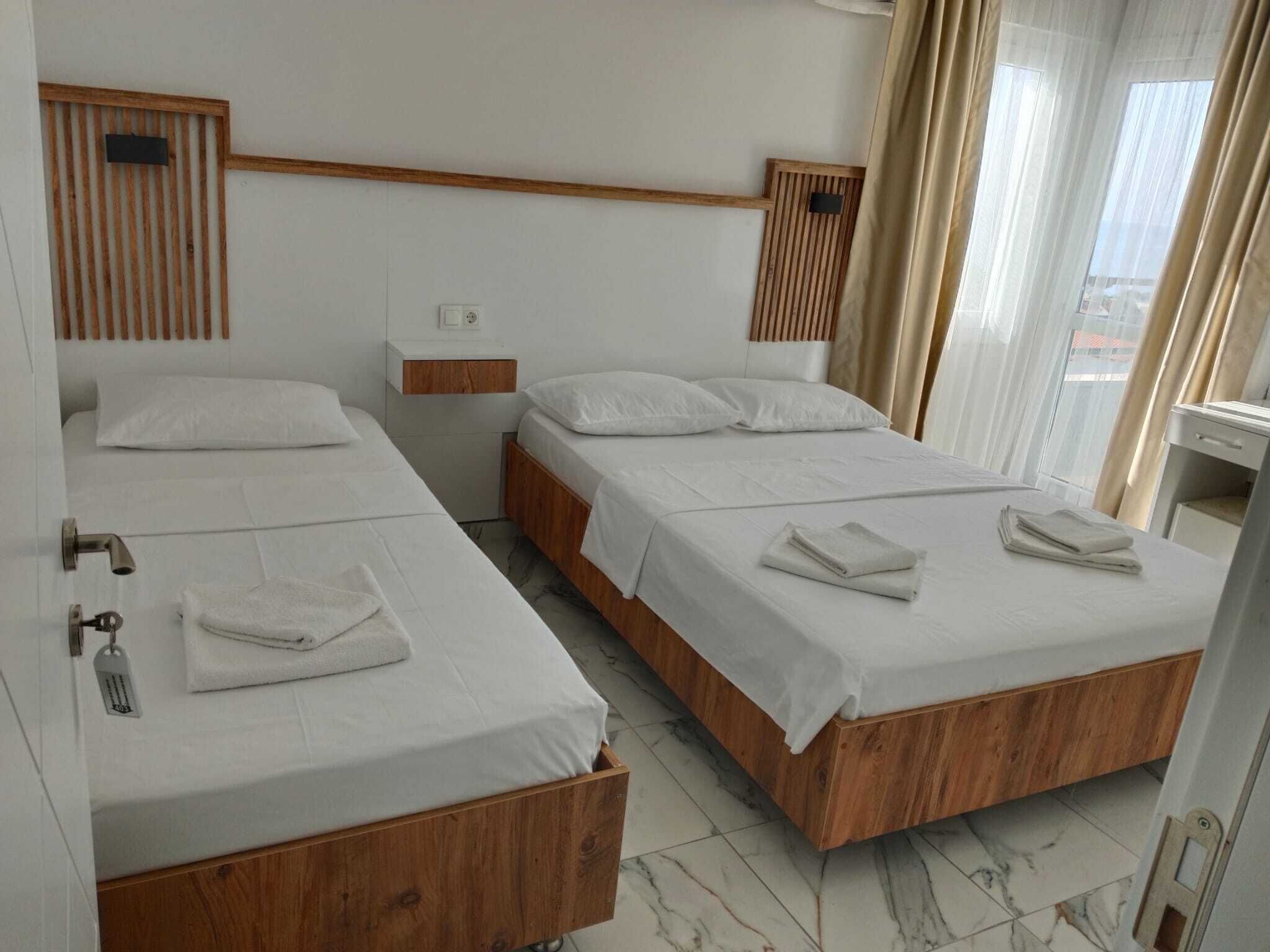 Ein Hotelzimmer mit zwei Einzelbetten, Holz-Kopfteilen, einem kleinen Nachttisch und einem Fenster mit Vorhängen sowie Blick ins Freie.