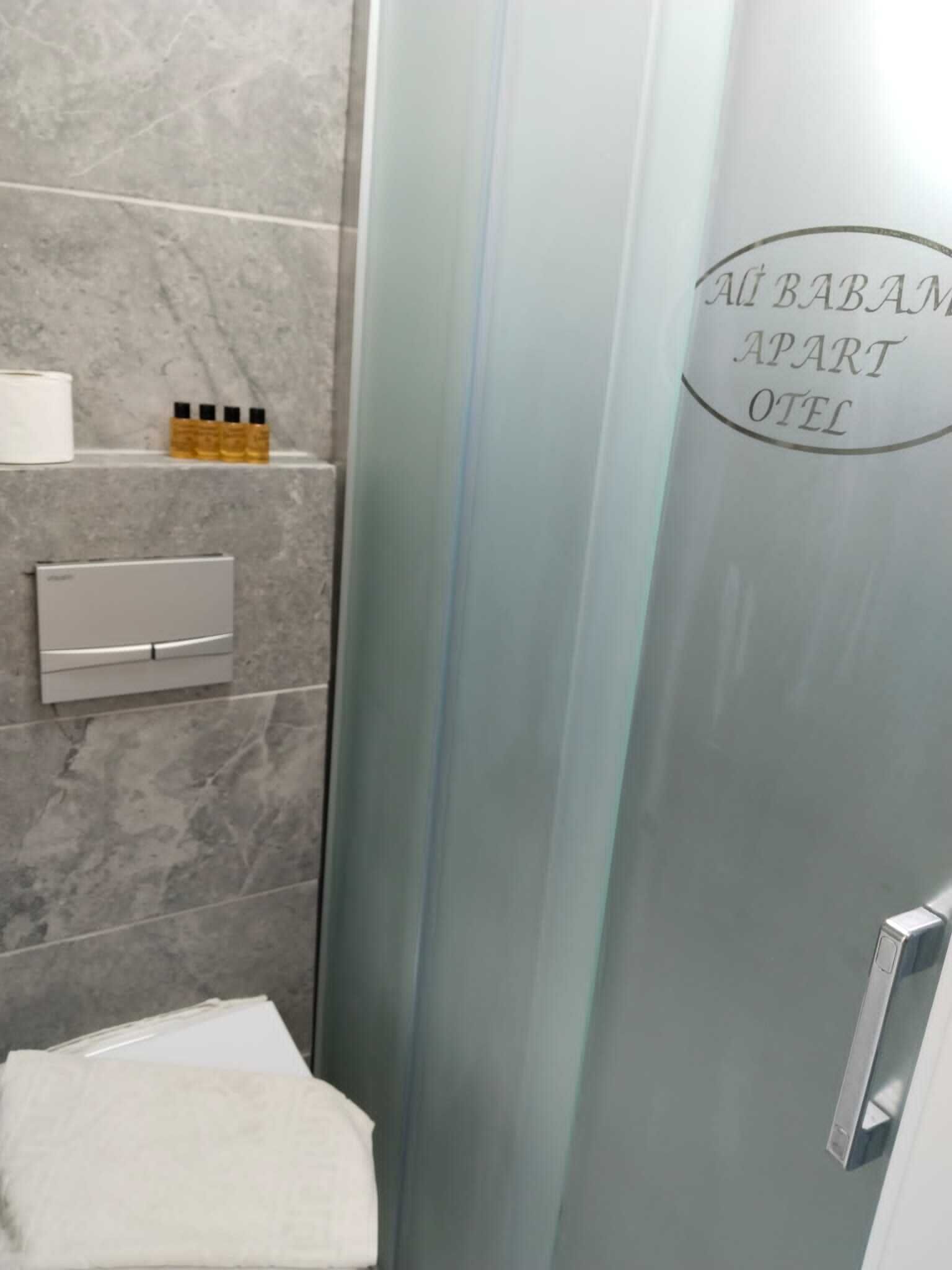 A bathroom with a glass shower enclosure, a toilet, and a towel rack. The shower has a logo that reads "AÜ BABAM APART OTEL".