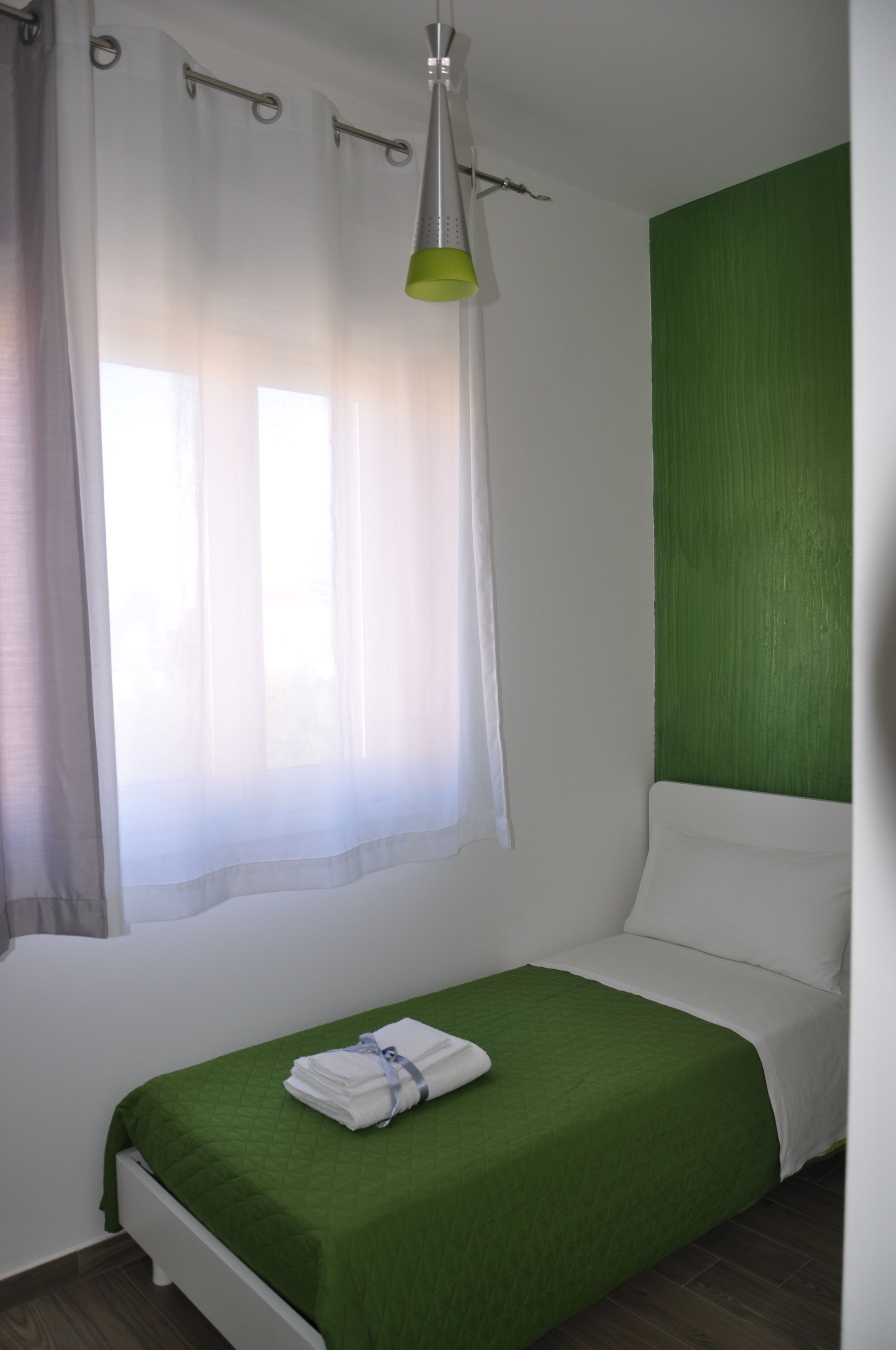 A single bed with a green bedspread, white pillows, and a neatly folded white towel on the side.