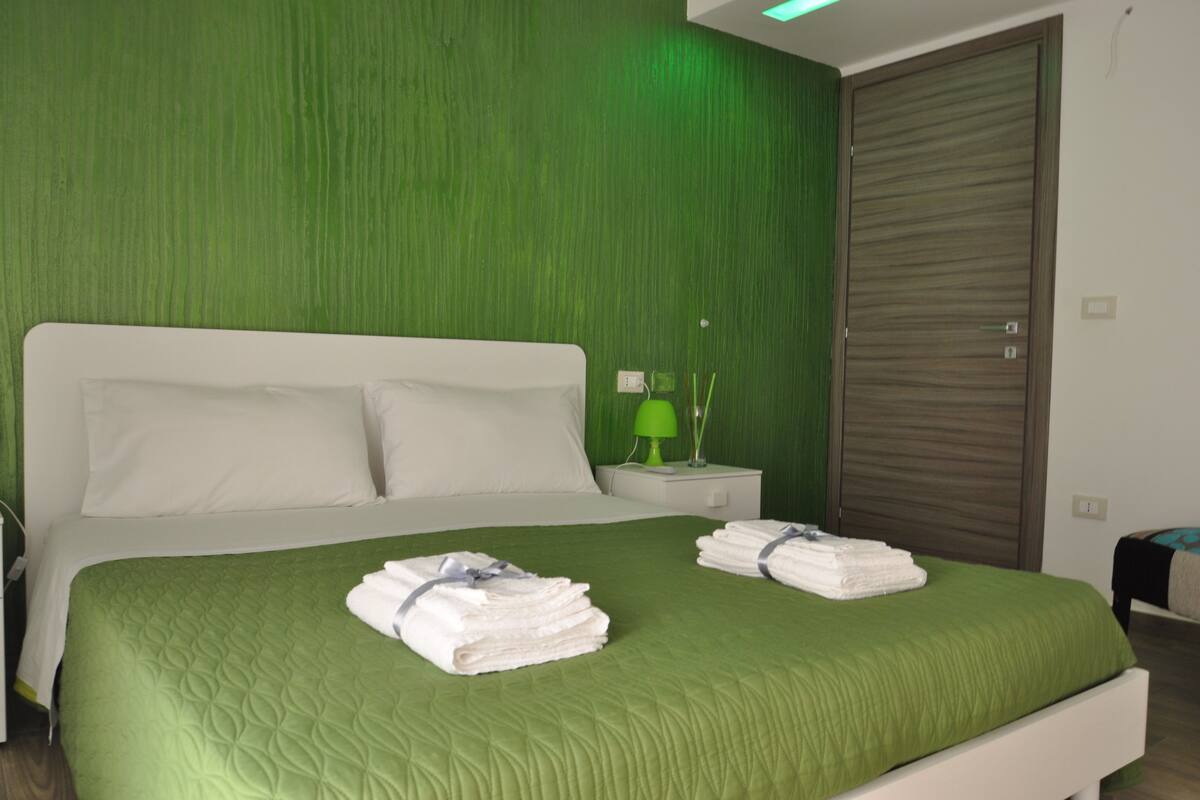A neatly made double bed with green bedding, a bedside table with a lamp, and a closed wardrobe.
