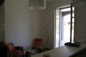 Quarto com escrivaninha branca, cadeiras coloridas, luminária pendente e porta que dá para o exterior.