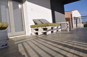 A balcony with a wooden floor, a white wall, and a grey cushion on a wooden bench.