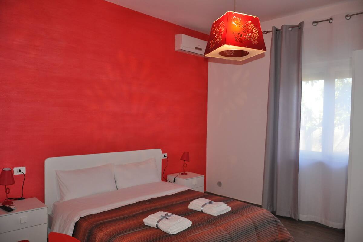 A hotel room with a red wall, a white bed, a red lamp, and a window with curtains.