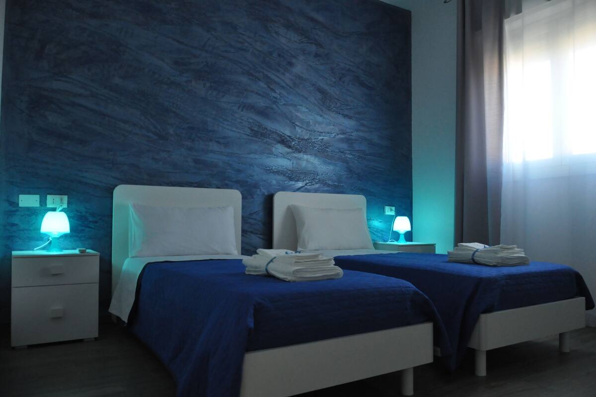 A hotel room with two beds, each with a blue bedspread and white pillows. There are two bedside tables with lamps, and a window with curtains.
