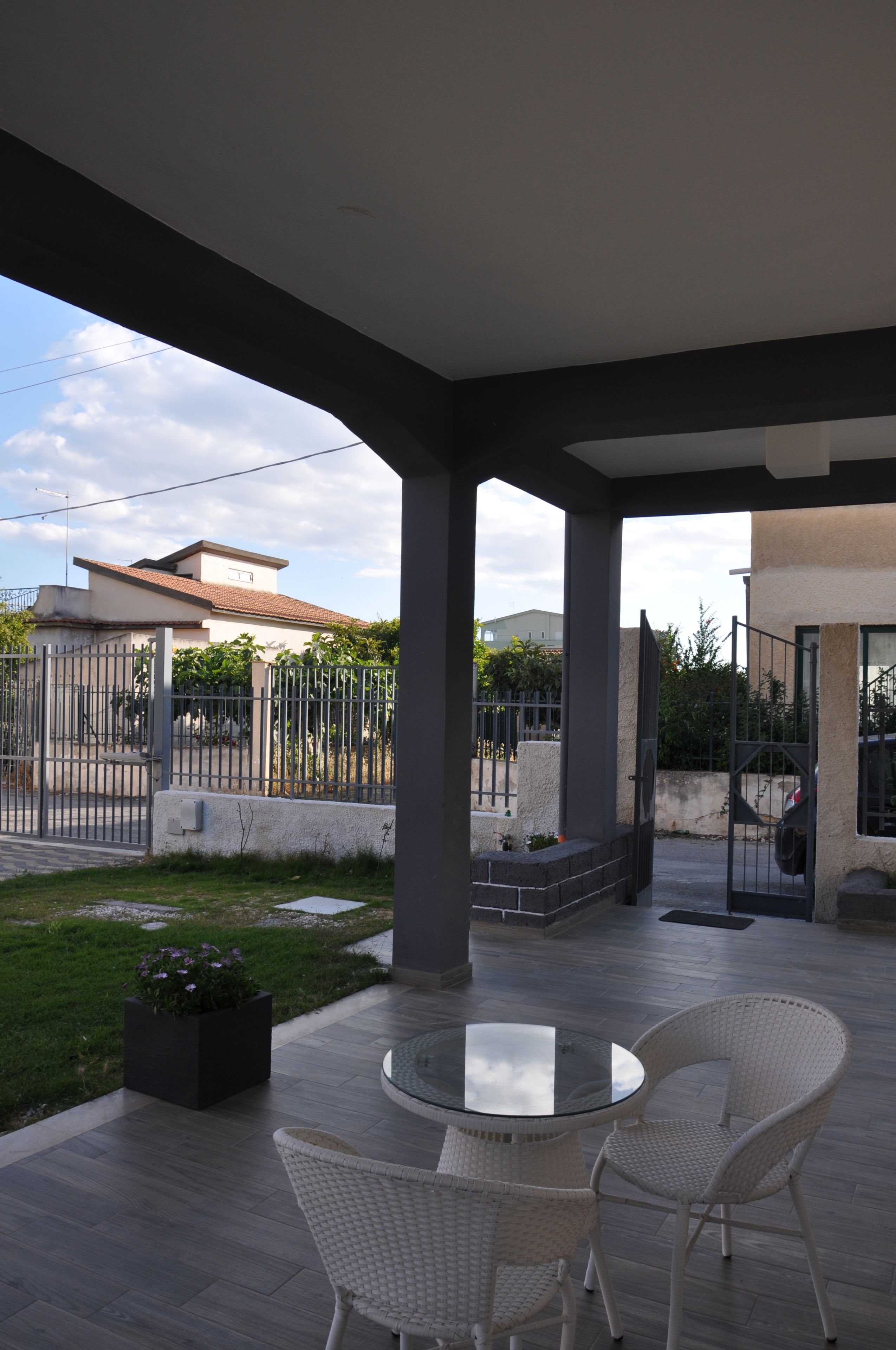 Une terrasse couverte avec une table ronde en verre et deux chaises en osier, un bac à fleurs en béton agrémenté de plantes, et une vue sur une maison et un portail au fond.