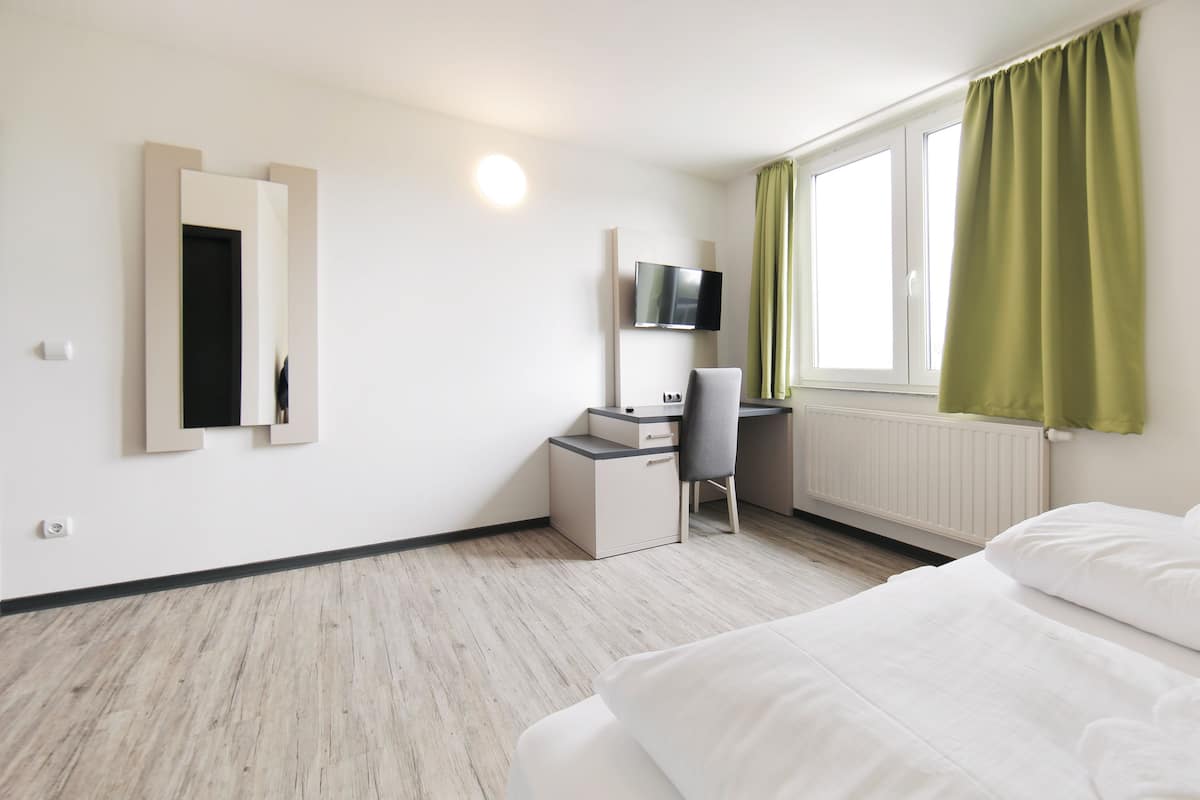 Een hotelkamer met een bed, een bureau met stoel, een aan de muur bevestigde televisie en een raam met gordijnen.