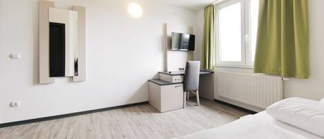 A hotel room with a bed, a desk with a chair, a TV mounted on the wall, and a window with curtains.