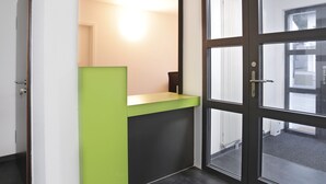 Een receptiebalie met een groen aanrecht en een zwart onderstel, een glazen deur met een hendel, en een radiator zichtbaar door het glas.