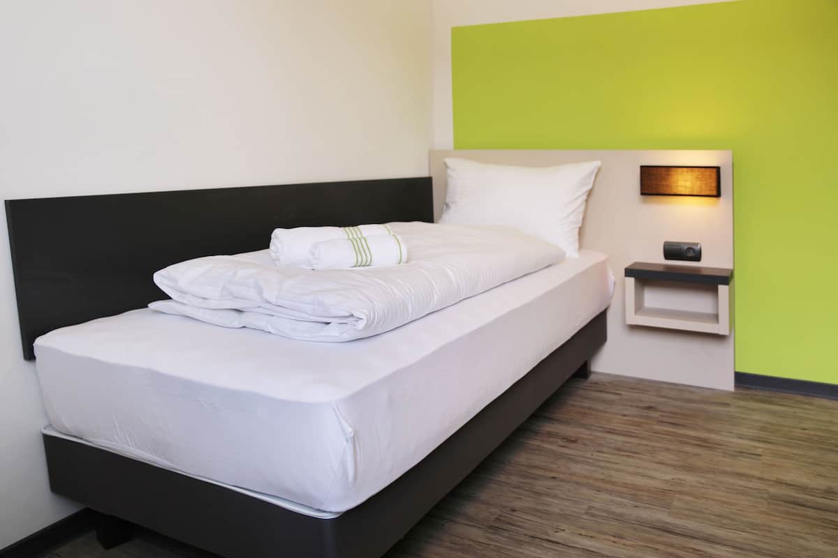 Een netjes opgemaakt bed met witte lakens en een opgevouwen handdoek, een zwart hoofdbord, een nachtkastje met een lamp en een groene accentmuur.