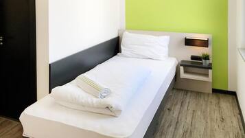 A neatly made bed with white linens and a rolled towel, a bedside table with a lamp and a small plant, and a green accent wall.