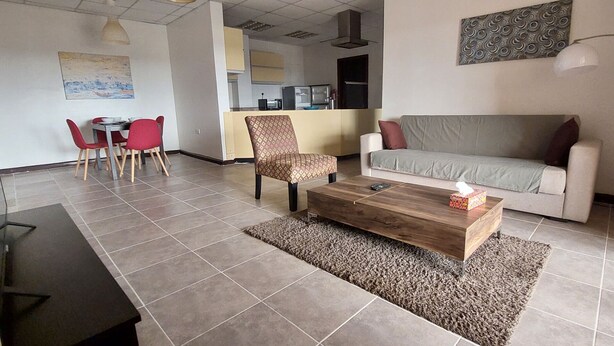 A living room with a sofa, a coffee table, and a patterned armchair. There is a dining area with red chairs and a table in the background.