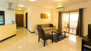 A modern living room with a sofa, a chair, a coffee table, and a TV. There is a kitchen area with a counter and a sink. The room has a balcony with a view.