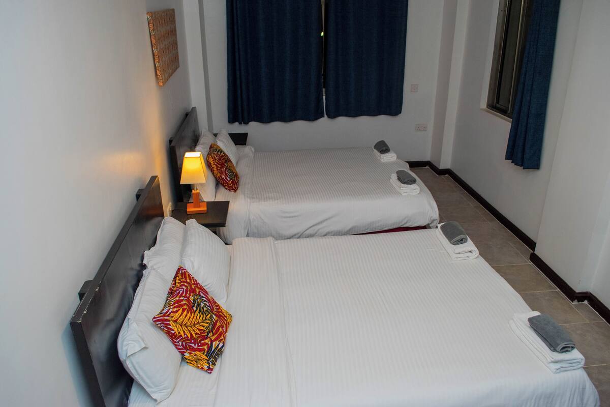 Una habitación de hotel con dos camas, una mesita de noche con lámpara y una ventana con cortinas.