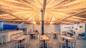 A spacious indoor area with wooden ceiling beams, multiple tables and chairs, a bar area, and a visible kitchen. - Aelia Apartments (Hersonissos)