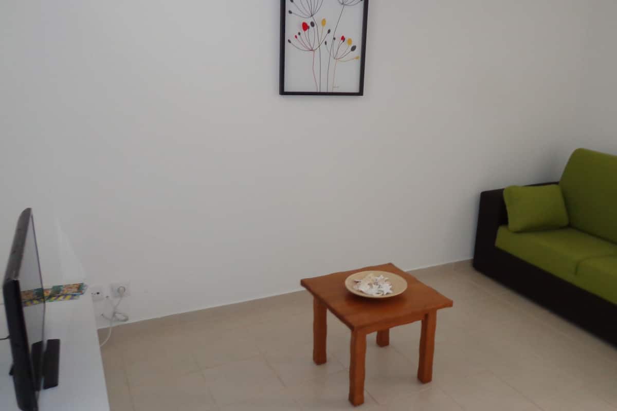 Un salon avec un canapé vert, une petite table en bois sur laquelle se trouve une assiette, et un tableau encadré accroché au mur.