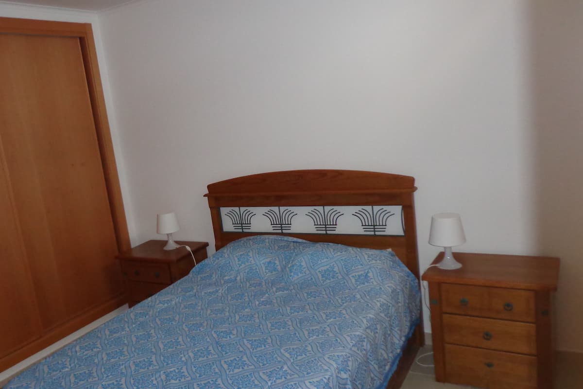 Une chambre à coucher avec un lit en bois, deux tables de chevet avec des lampes, une armoire en bois et une couvre-lit à motifs.