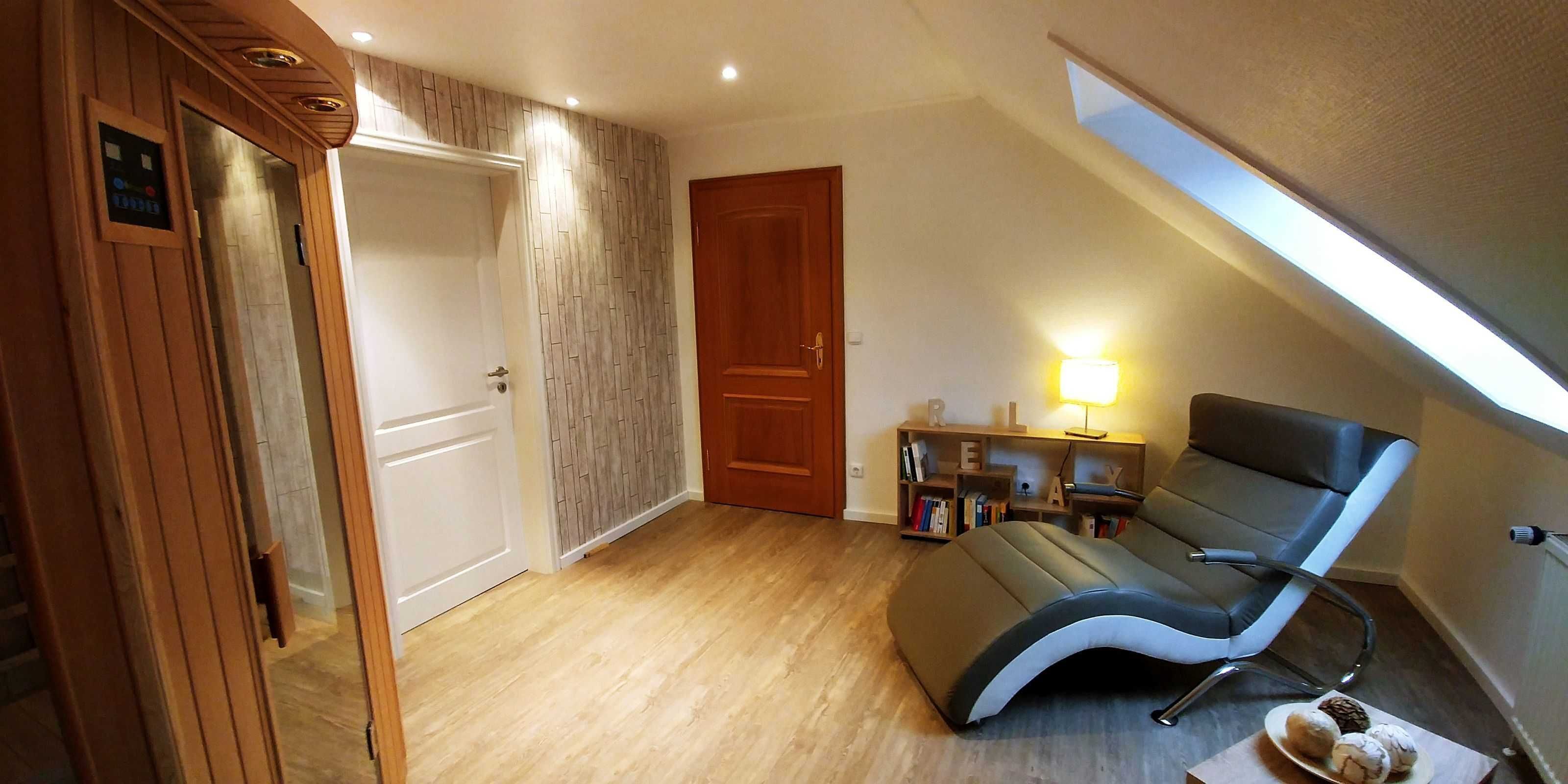 Una habitación con piso de madera, una silla reclinable, una estantería llena de libros, una lámpara y una puerta.