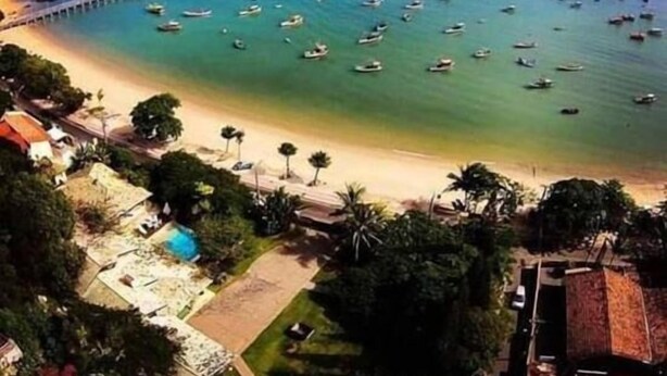 A coastal area with numerous boats on the water, a pier, and a beach with buildings and greenery. - Encanto Familiar (Penha)