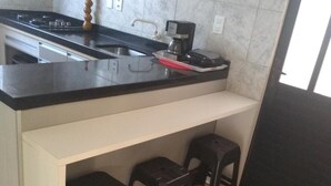 A compact kitchen with a black countertop, a coffee machine, and a small sink. There are three stools under the counter. - Encanto Familiar (Penha)