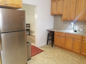 A kitchen with wooden cabinets, a stainless steel refrigerator, a granite countertop, and a small stool.