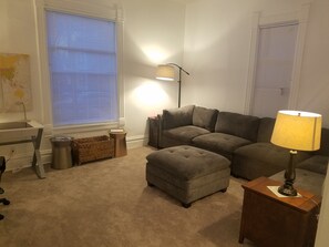 A living room with a grey sofa, a small ottoman, a lamp, a globe on a shelf, and a window with blinds.