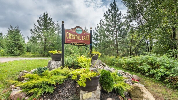 A sign for Crystal Lake with a welcome message, surrounded by a landscaped garden with various plants and rocks. - Crystal Palace Cottage, Kawartha Lakes (Kinmount)