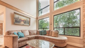 A spacious living room with a large sectional sofa, a glass coffee table, and a large window offering a view of trees. - Crystal Palace Cottage, Kawartha Lakes (Kinmount)