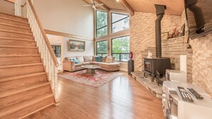 A spacious living room with a wooden staircase, a stone fireplace, and large windows. - Crystal Palace Cottage, Kawartha Lakes (Kinmount)
