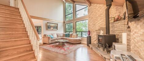 A spacious living room with a wooden staircase, a stone fireplace, and large windows.