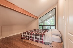 A bedroom with a large window, a wooden ceiling beam, and a plaid bedspread. - Crystal Palace Cottage, Kawartha Lakes (Kinmount)