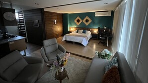 A modern hotel room with a bed, a desk, a chair, and a sofa. There is a clock on the wall and an air conditioning unit on the wall.