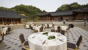 Een traditionele Koreaanse feestzaal met ronde tafels gedekt voor een diner, omgeven door houten gebouwen en een met stenen bestraat erf.