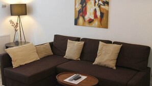 Un sofá seccional marrón con cojines beige, una pequeña mesa redonda de madera con un control remoto y una lámpara de pie.