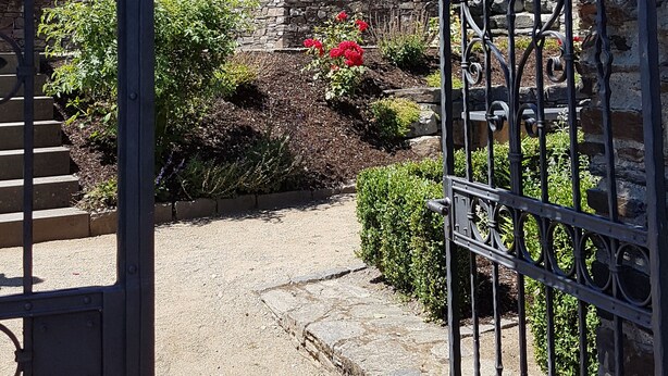Un edificio de piedra con una torre, una reja de metal y un jardín con flores rojas.