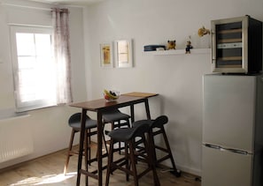 Eine kleine Küche mit einem Holztisch, zwei Barhocker, einem Kühlschrank und einem Fenster mit Vorhängen.