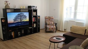 Una sala de estar con una televisión de pantalla plana, una estantería, un sillón estampado, un sofá marrón y una mesita con una lámpara.