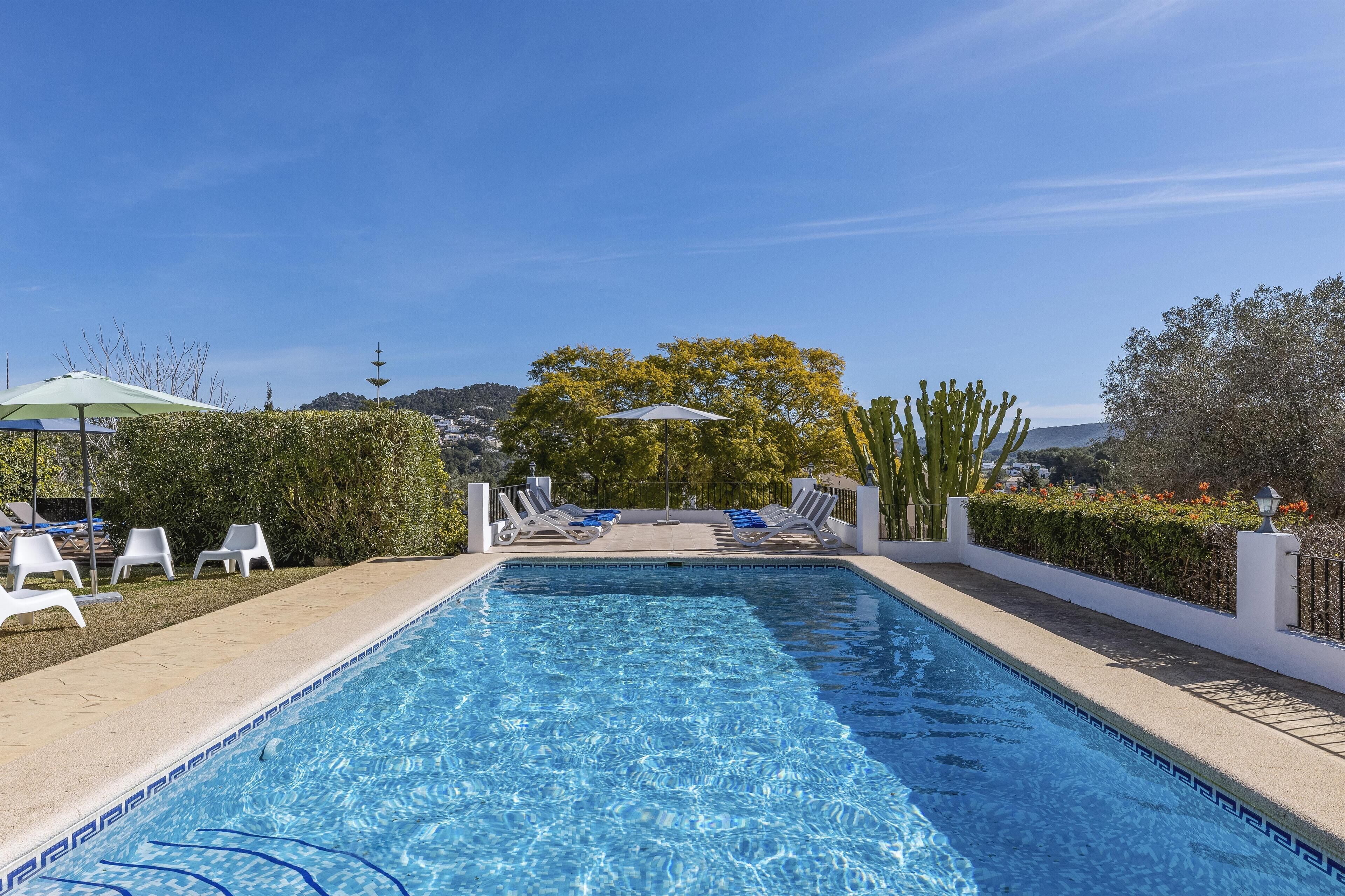 A rectangular swimming pool with clear blue water, surrounded by white lounge chairs and umbrellas, with a view of trees and hills in the background.