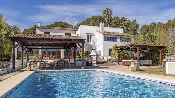 A swimming pool with a pergola and outdoor seating area, surrounded by a white building with a tiled roof and a garden.