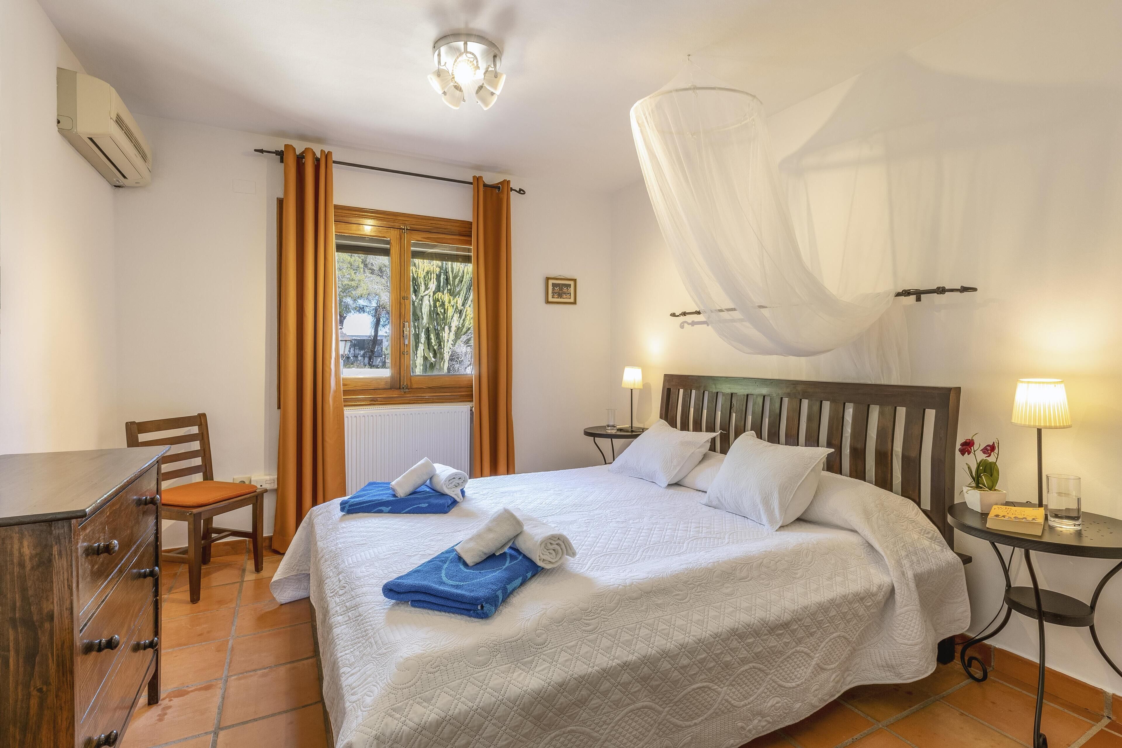 A hotel room with a bed, a desk, a chair, a window with curtains, a bedside table with a lamp, and a wall-mounted air conditioner.